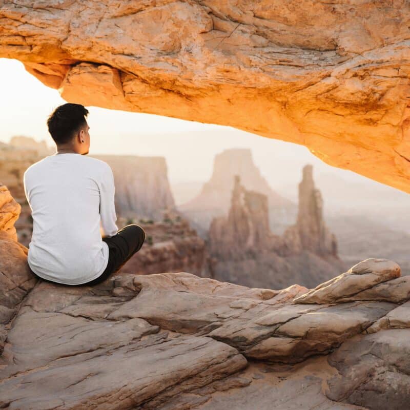 Man sitting on a rocky ledge in a desert canyon, with sandstone formations and distant arches in warm evening light