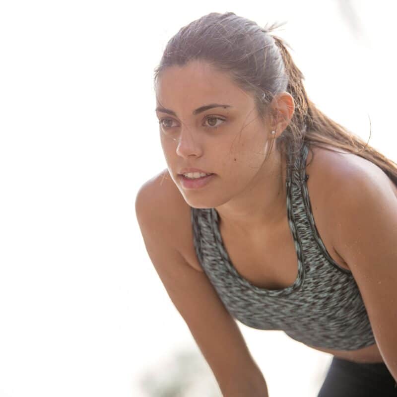 Woman in a gray patterned sports bra and black leggings leaning forward in a bright outdoor setting, performing a stretch.