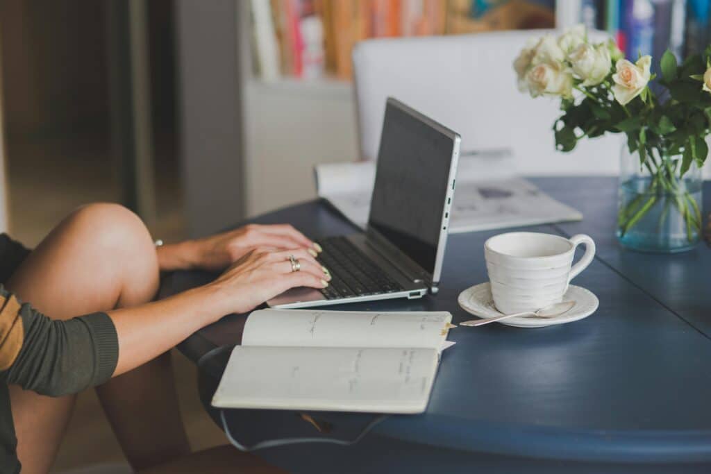 pexels-peter-olexa-2214257-4012966 - Vintage at Tabernacle Person typing on a laptop at a desk, with an open notebook, a white mug, and a vase of pale roses nearby.