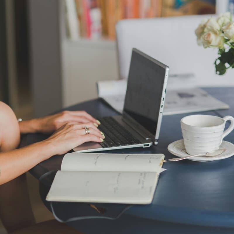 Person typing on a laptop at a desk, with an open notebook, a white mug, and a vase of pale roses nearby.